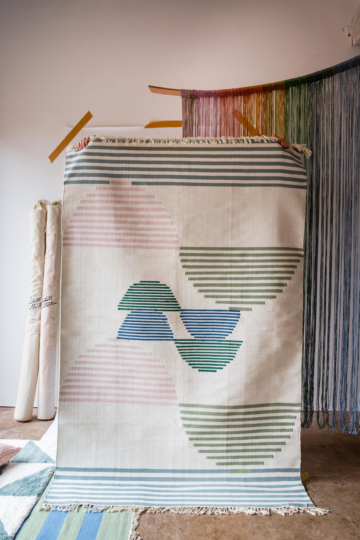 A person holding up a rug with an abstract stripe pattern. The background of the rug is cream and the pattern is in green, pink and blue